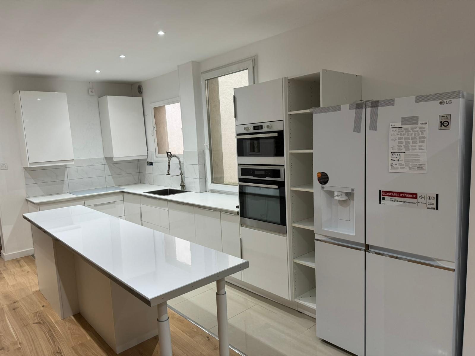Cuisine blanche brillante en cours de pose avec îlot central : entre la colonne des fours et le réfrigérateur, un caisson standard encore sans façade, tablettes apparentes