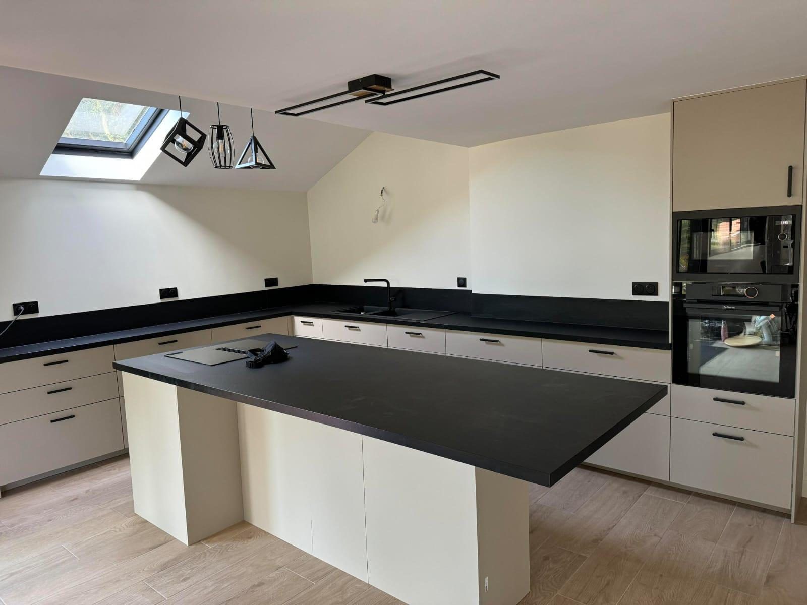 Cuisine achevée sous rampant : façades claires, plan de travail sombre, îlot central et fenêtre de toit. La gamme des fournitures retenue pèse à elle seule une part de l'écart entre deux chiffrages. Chantier réel des équipes partenaires.