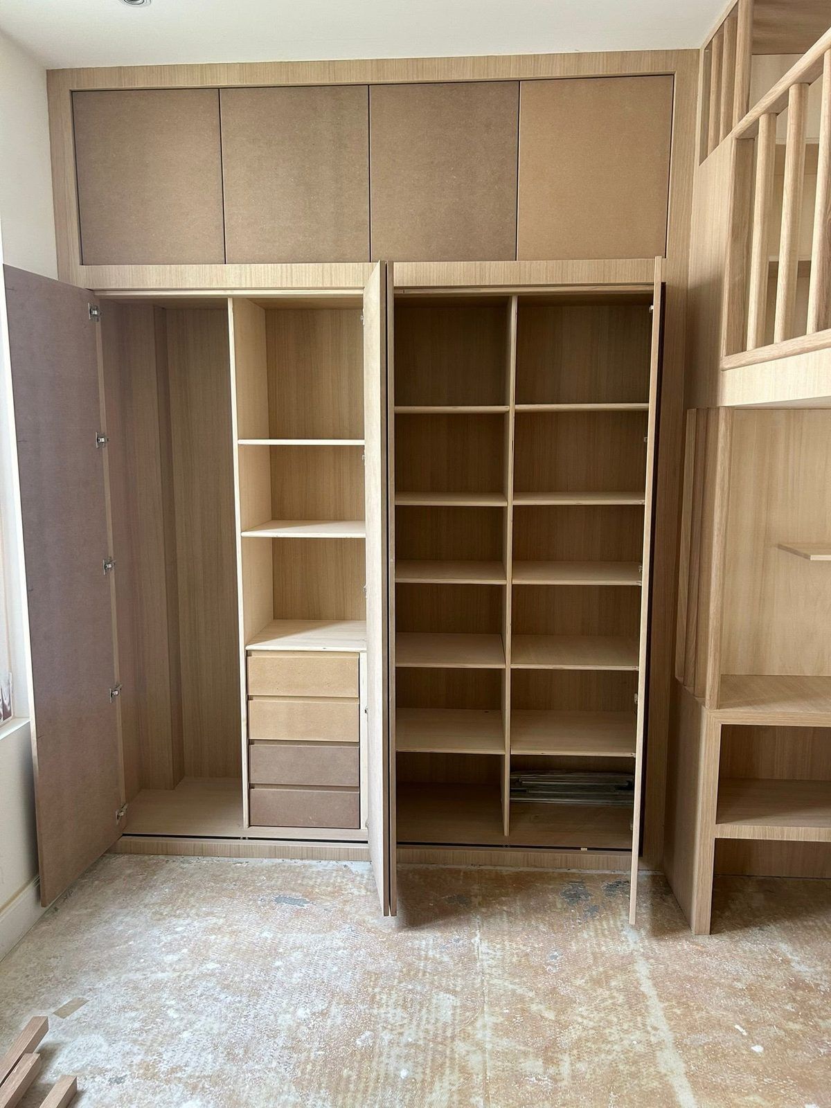 Deux ouvrages de menuiserie dans la même pièce : des rangements en panneau brut, portes ouvertes sur leurs tablettes, et à droite l'escalier bois avec ses marches et son garde-corps à barreaux. Chantier réel des équipes partenaires.