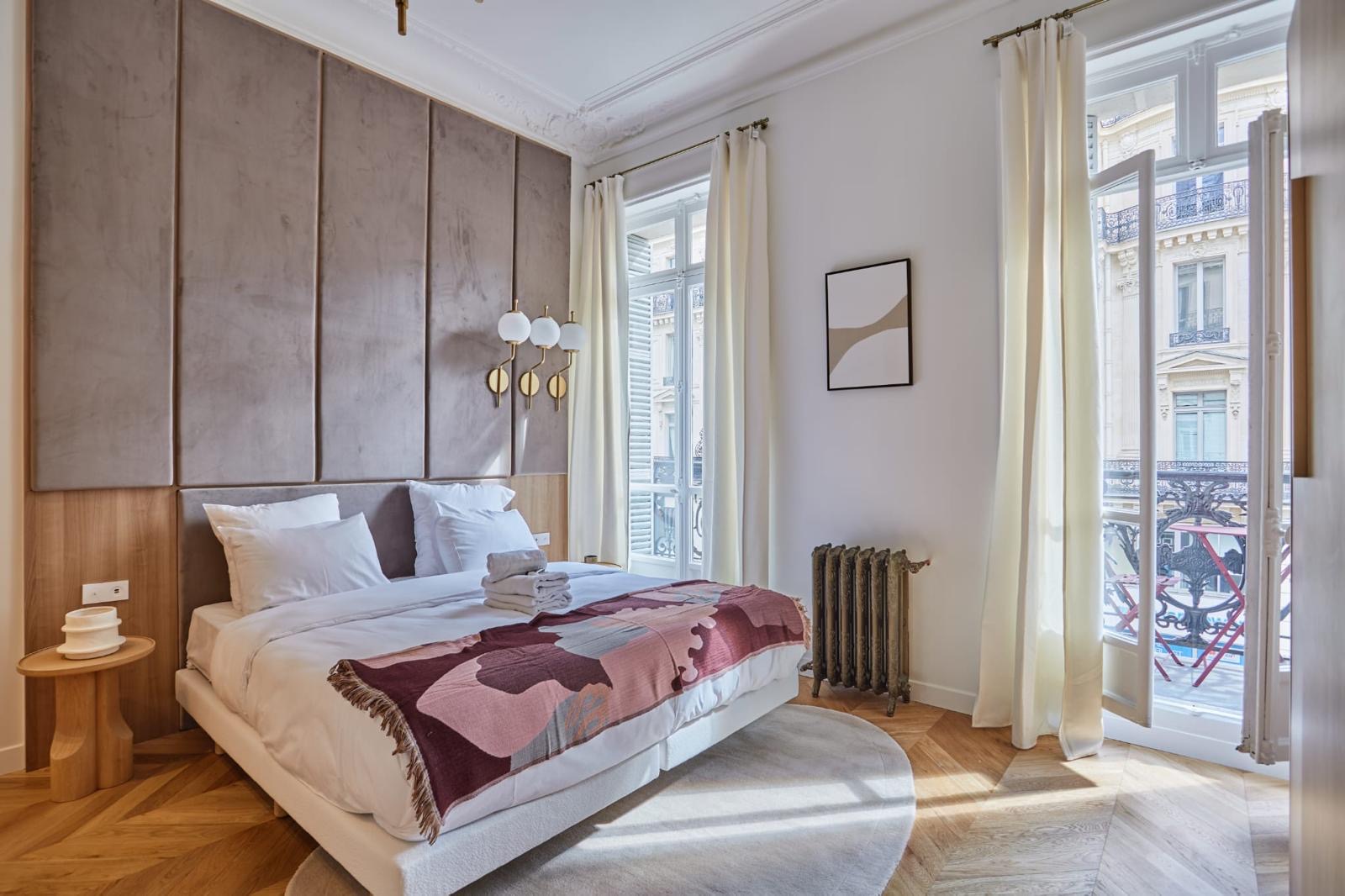 Chambre sur rue rénovée : tête de lit en panneaux capitonnés taupe, radiateur en fonte conservé, parquet chêne à chevrons