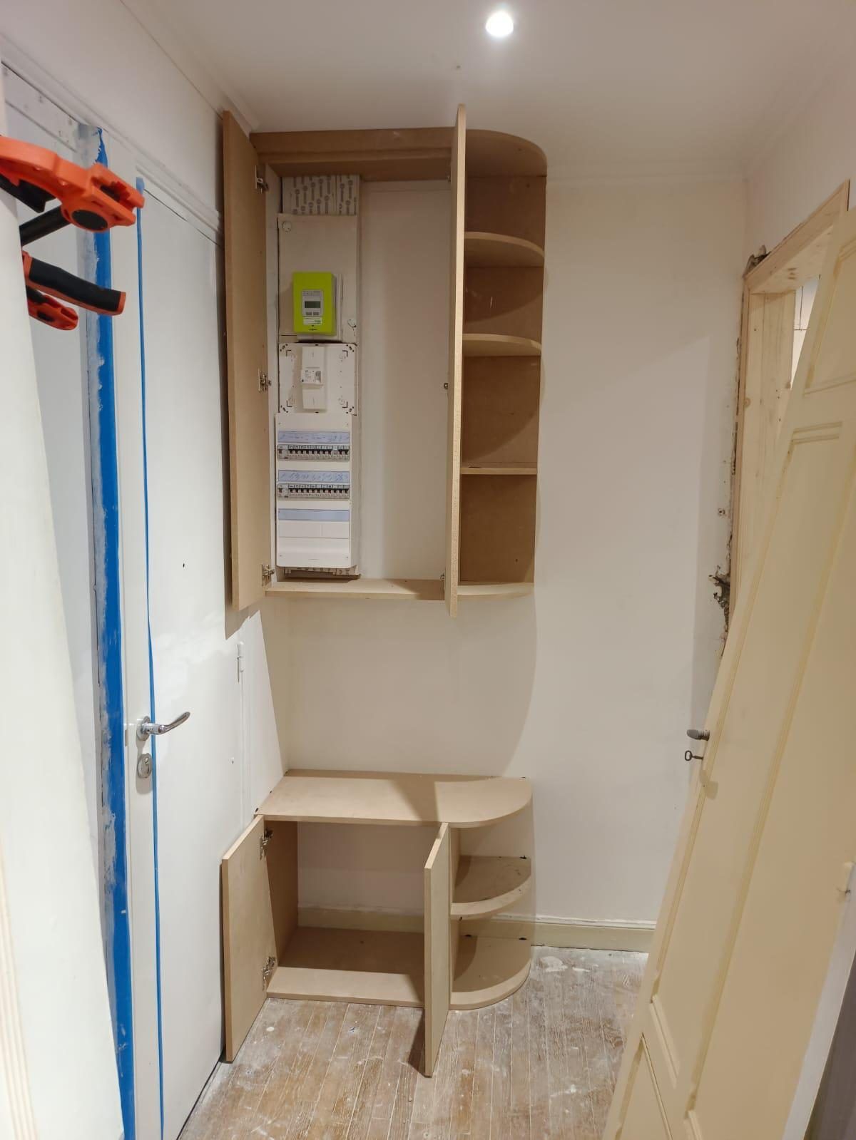 Meuble d'entrée refermant le compteur et le tableau électrique, portes ouvertes en cours de pose : l'accès aux organes de coupe se vérifie avant que la menuiserie ne le referme. Chantier réel des équipes partenaires.