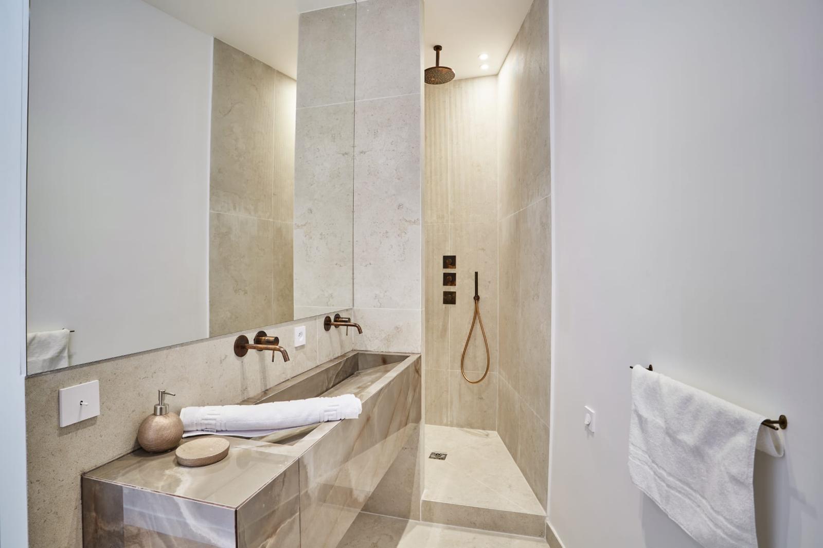Salle d'eau rénovée : douche à l'italienne de plain-pied en carrelage ton pierre claire, siphon de sol, ciel de pluie et robinetterie encastrée cuivrés, plan-vasque en pierre veinée